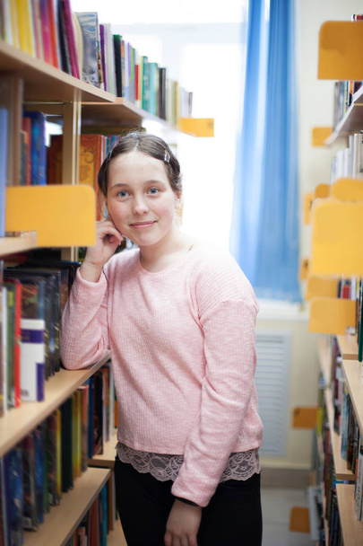 Schattig tiener meisje in openbare bibliotheek in de buurt van planken met boeken. - Foto, afbeelding