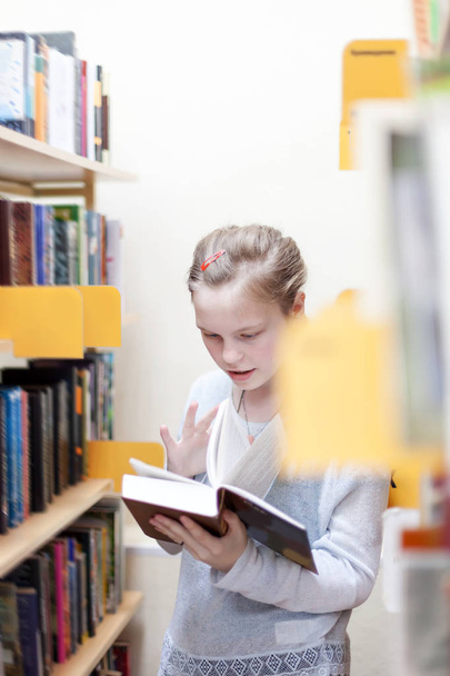 Χαριτωμένο έφηβος κορίτσι σε δημόσια βιβλιοθήκη κοντά ράφια με βιβλία. - Φωτογραφία, εικόνα