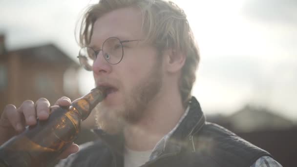 Portrait of the blond man drinking beer outdoors looking away. Bearded man in glasses enjoying his alcohol drink, hair fluttering in the wind. Slow motion - Video, Çekim