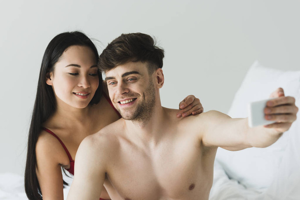 handsome shirtless man making selfie with smartphone near attractive asian girlfriend  - Photo, image