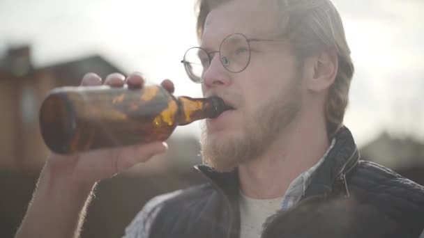 Bearded man with glasses drinking beer and enjoying beverage outdoors. Guy tastes lager from bottle. Slow motion. - Video, Çekim