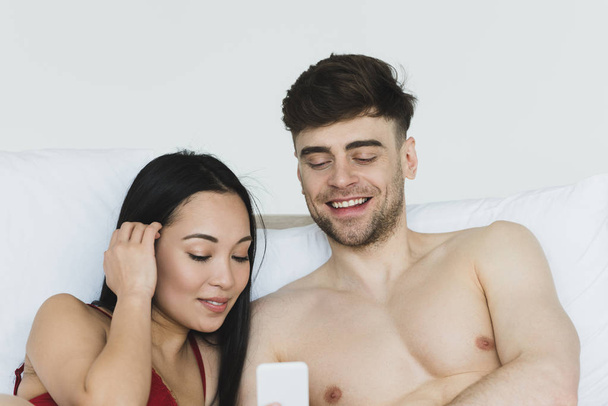 szép ázsiai nő piros fehérnemű segítségével okostelefon félmeztelen barátja az ágyban - Fotó, kép