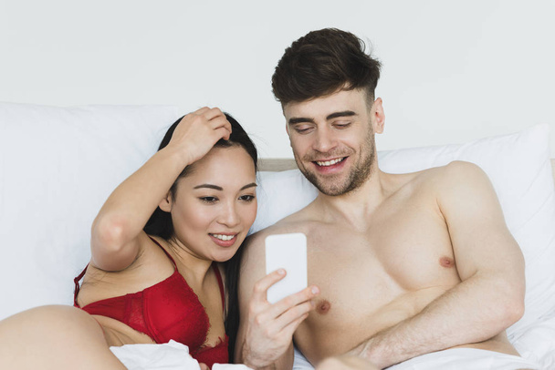 handsome man using smartphone while lying in bed near pretty asian girlfriend in red lingerie - Φωτογραφία, εικόνα