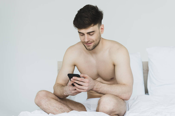 красивий, усміхнений чоловік в нижній білизні сидить у ліжку і використовує смартфон
 - Фото, зображення