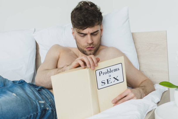 ernste hemdlose Mann in blauen Jeans lesen Probleme in Sex-Buch im Bett liegend - Foto, Bild