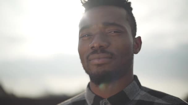 Portrait of handsome african american man looking into the camera outdoors. The sunny light sky in the background. Slow motion. - Filmagem, Vídeo