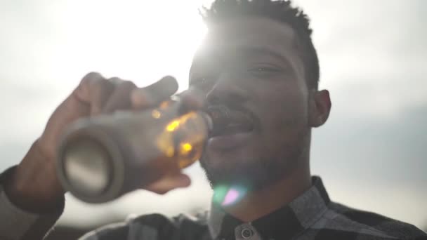 Retrato de um homem afro-americano bonito a beber cerveja a olhar para a câmara ao ar livre. O céu ensolarado ao fundo. Jovem que gosta da bebida alcoólica. Movimento lento
. - Filmagem, Vídeo