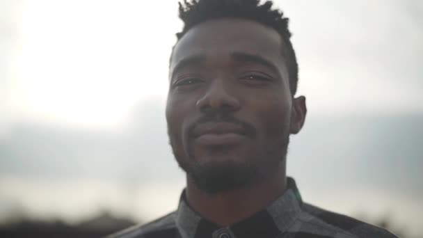 Close up portrait of handsome african american man looking into the camera outdoors. The sunny light sky in the background. Slow motion. - Filmagem, Vídeo