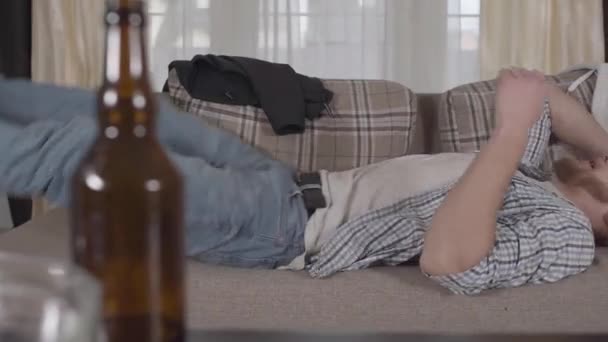 Bearded man in casual clothes falls on the sofa and throws glasses on the table. Beer bottle in the foreground. There is mess in the apartment. Unhealthy lifestyle. Slow motion - Imágenes, Vídeo