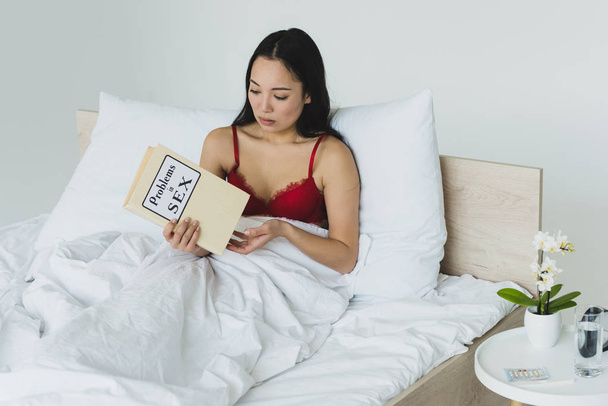 задумлива азіатська жінка в червоній білизні, що тримає проблеми в секс-книзі, лежачи в ліжку
 - Фото, зображення