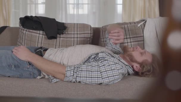 Bearded man in casual clothes lying on the sofa. Blurred bottle in the foreground. There is mess in the apartment. Unhealthy lifestyle, problem with alcohol. Slow motion - Materiał filmowy, wideo
