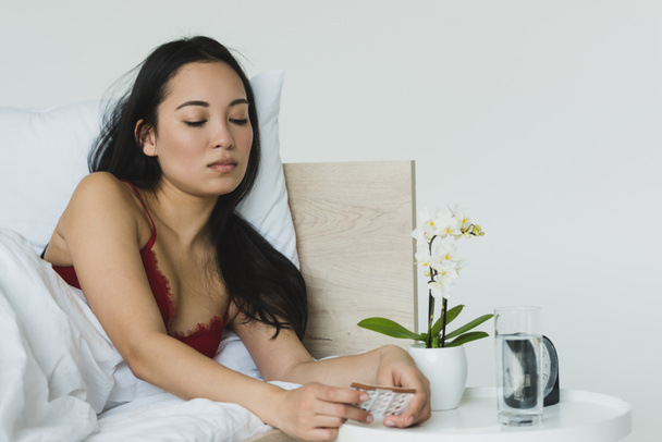 mooie, trieste Aziatische vrouw met geboortebeperking pillen terwijl liggend in bed in de buurt van de tafel met orchidee in bloempot - Foto, afbeelding