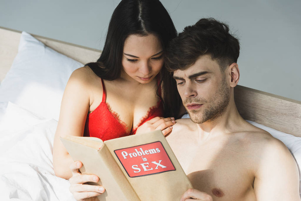 thoughtful multicultural couple reading problems in sex book while lying in bed  - Foto, Imagem