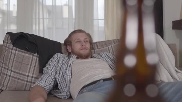 Young bearded guy falls asleep on the couch. Alcoholic beverages are on the table before the bed. - Materiał filmowy, wideo