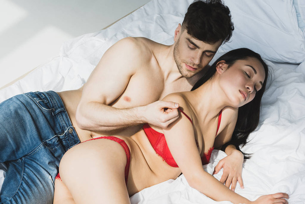 sexy shirtless man kissing passionate asian girlfriend in red lingerie while lying on white bedding - Foto, Imagen