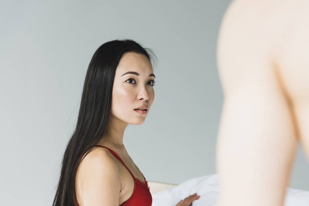 nachdenkliche, schöne asiatische Frau in rotem BH schaut ihren hemdlosen Freund an  - Foto, Bild