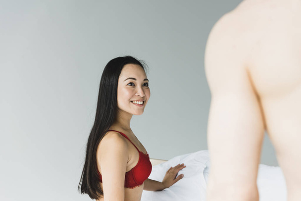 attractive, smiling asian woman in red lingerie looking at shirtless boyfriend in bedroom - Фото, зображення