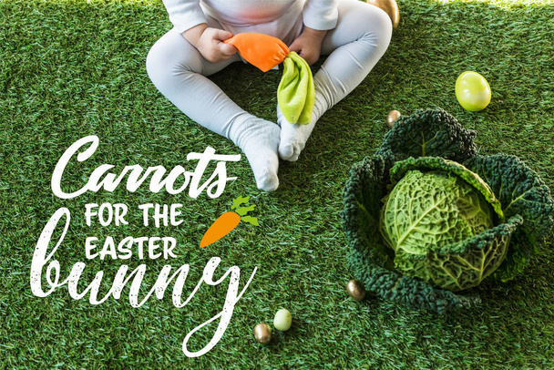 partial view of child holding toy carrot while sitting near Easter eggs and savoy cabbage on green grass with carrots for the Easter bunny lettering - Photo, Image