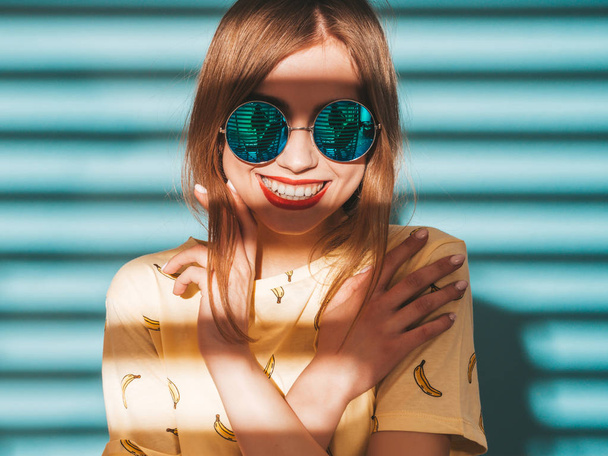 Μια νεαρή όμορφη γυναίκα που κοιτάζει την κάμερα. Μοντέρνο κορίτσι σε casual καλοκαιρινά κίτρινα ρούχα T-shirt. Θετικό θηλυκό δείχνει συναισθήματα προσώπου σε γυαλιά ηλίου. Αστείο μοντέλο ποζάρει κοντά σε μπλε τοίχο - Φωτογραφία, εικόνα