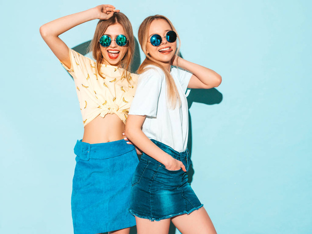 Two young beautiful smiling blond hipster girls in trendy summer jeans skirts clothes. Sexy carefree women posing near blue wall in round sunglasses. Positive models having fun and showing peace sign - Fotoğraf, Görsel