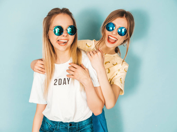Two young beautiful smiling blond hipster girls in trendy summer colorful  clothes. Sexy carefree women posing near blue wall in round sunglasses. Positive models having fun - Фото, зображення
