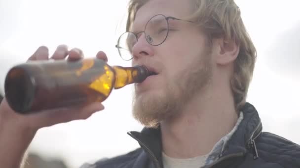 Bearded man with glasses drinking beer and enjoying beverage outdoors. Guy tastes lager from bottle. - Video, Çekim