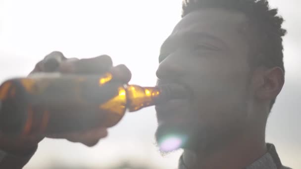 Portrait of handsome african american man drinking beer looking into the camera outdoors. The sunny light sky in the background. Young man enjoying his alcohol drink. - Imágenes, Vídeo