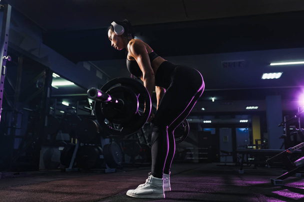 Attractive fit sexy woman in wireless headphones listen music and doing crouches with a barbell in the gym. Woman training back - Photo, image
