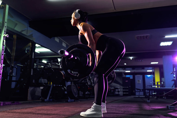 Attractive fit sexy woman in wireless headphones listen music and doing crouches with a barbell in the gym. Woman training back - Photo, Image