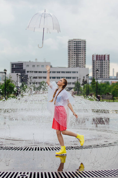 Fiatal csinos lány, két zsinór sárga csizma dobás és elfogást átlátszó esernyő szökőkút közelében. - Fotó, kép