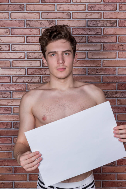 Young naked sexy guy posing in underwear with a billboard on the background of a brick wall - Photo, Image