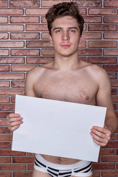 Young naked sexy guy posing in underwear with a billboard on the background of a brick wall - Zdjęcie, obraz