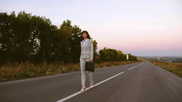 mulher de negócios com pasta preta caminha em terno leve e sapatos brancos de salto alto sai da cidade ao longo do asfalto com marcas brancas
 - Foto, Imagem