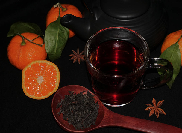 Tè d'autunno in una tazza trasparente. Tagli tangerini su uno sfondo nero. Tè in un cucchiaio di legno
 - Foto, immagini