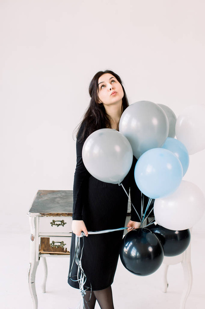 Beautiful young woman with balloons on a white background, standing near the vintage white table and looking up. Birthday and party. Girl in black dress at the festival - Φωτογραφία, εικόνα