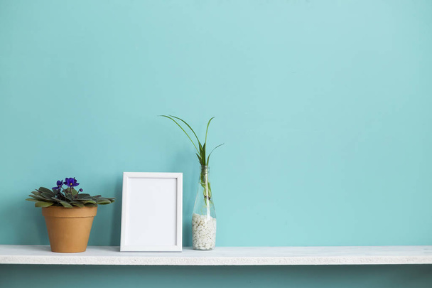 Moderne kamer decoratie met Picture Frame mockup. Witte plank tegen pastel Turquoise muur met spin plant stekken in water en violet. - Foto, afbeelding