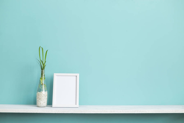 Moderna decoración de la habitación con marco de imagen maqueta. Estante blanco contra pared de color turquesa pastel con esquejes de plantas de araña en agua
. - Foto, imagen
