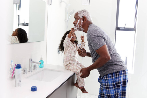 Grandfather Wearing Pajamas In Bathroom Shaving Whilst Granddaughter Watches - 写真・画像