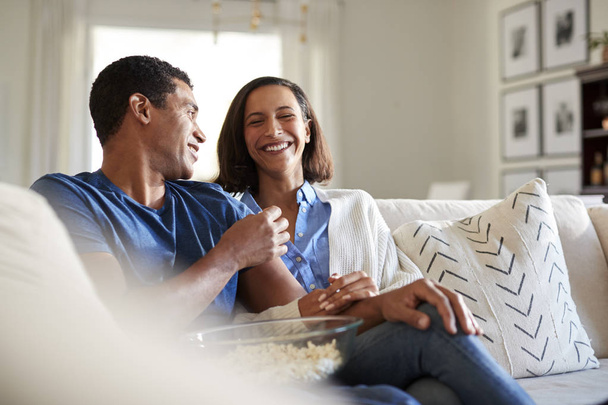 Young adult mixed race couple sitting on the sofa in their living room laughing and eating popcorn, selective focus - Photo, Image