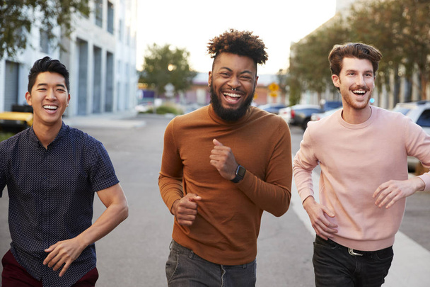Three hip young adult male friends running for fun in a city road, front view, close up - Foto, imagen