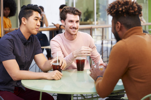 Three millennial male friends talking over cold brews outside a cafe in a city street, close up - Foto, afbeelding
