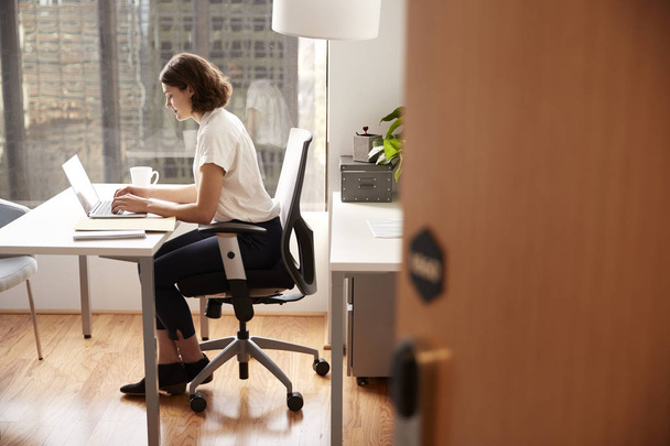 Businesswoman Sitting At Desk Working On Laptop In Modern Office Viewed Through Door Way - Fotó, kép