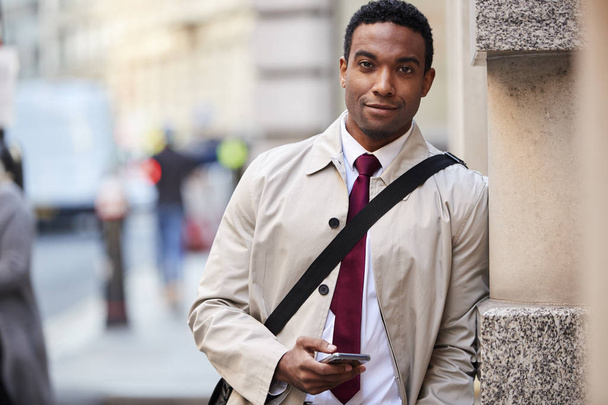 Young black businessman leaning on a wall in the street in London holding phone and smiling to camera, selective focus - Фото, изображение