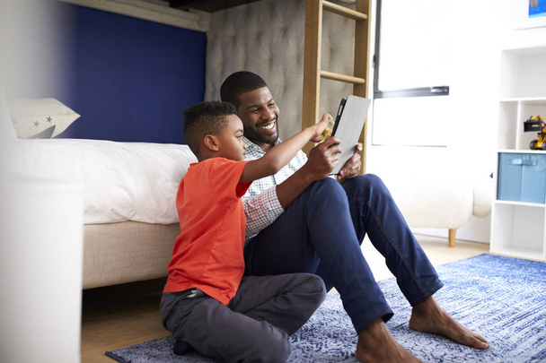 Father With Son Sitting In Bedroom Playing Game On Digital Tablet Together - Foto, imagen