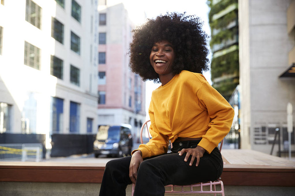 Young black woman with afro hair sitting on a chair in the street laughing to camera - Foto, Bild
