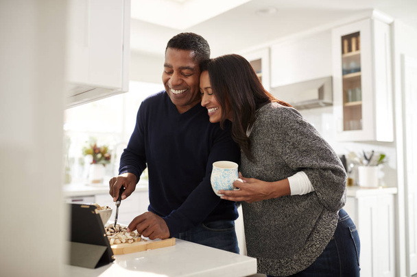 Middle aged mixed race couple standing at worktop in the kitchen preparing food following a recipe on a tablet computer, selective focus - Photo, Image