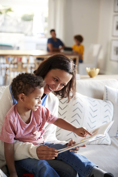 Close up of young mother sitting on a sofa in the living room reading a book with her toddler son, sitting on her knee, father and daughter sitting at a table in the background, vertical - Foto, Imagem
