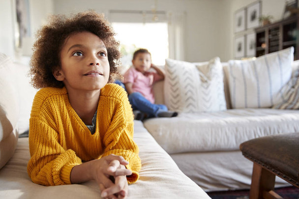 Close up of pre-teen girl lying on sofa watching TV in the living room, her younger brother sitting in the background, focus on foreground - Photo, Image