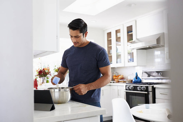 Millennial Hispanic man preparing cake mixture in kitchen, following a recipe on a tablet computer - Photo, Image