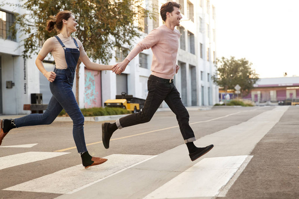 Millennial hipster couple running across the road in the city holding hands, side view - Photo, image